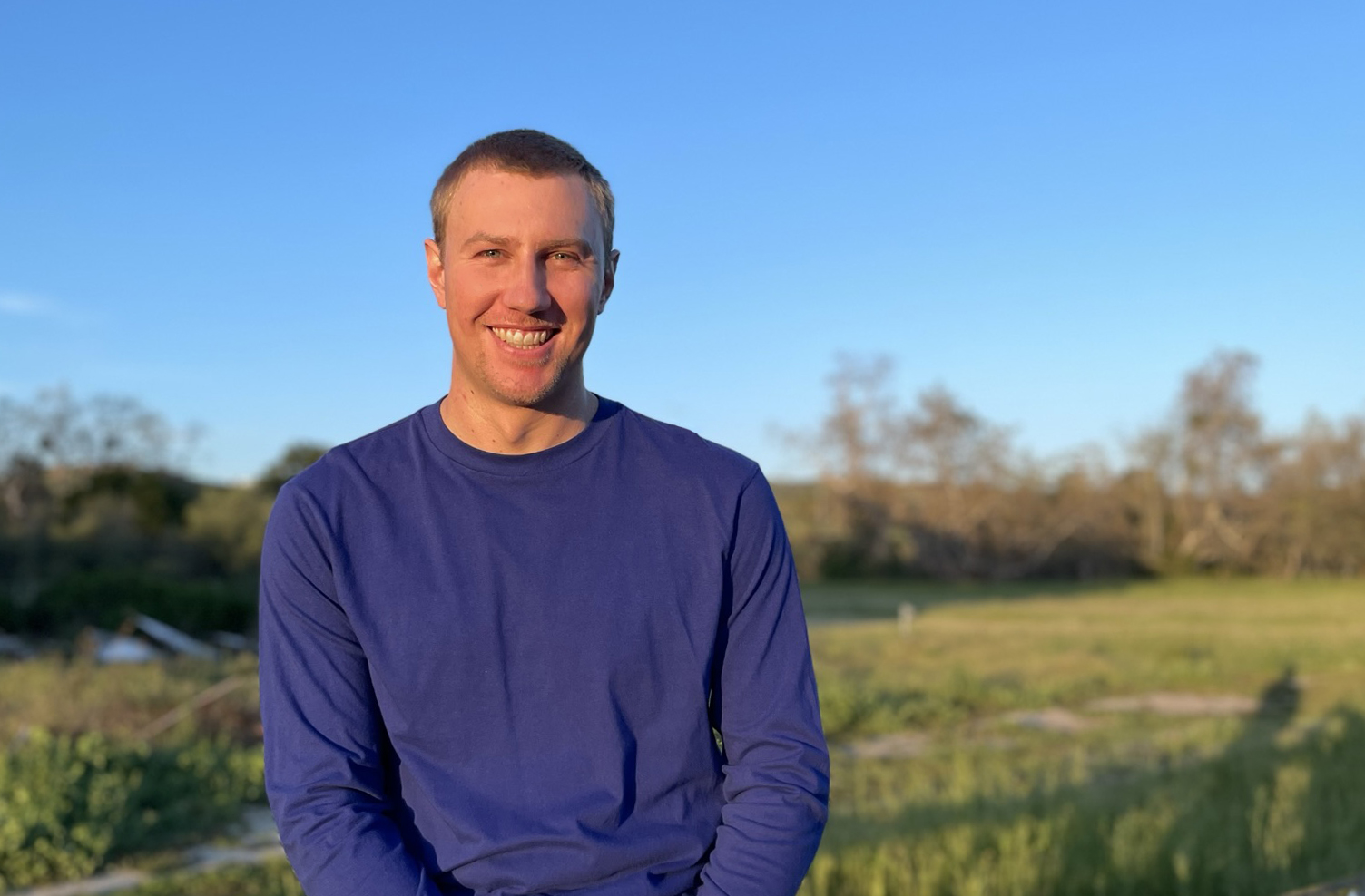 Chris smiling on a fence with grassy hilly background